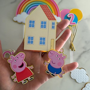 May include: Cake toppers featuring a rainbow, a house, and two pig characters. The house is beige with a red roof and blue window frames. The pig characters are pink, one in a red dress and the other in blue shorts. Includes a number 2 balloon.