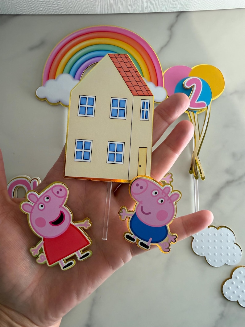 May include: Cake toppers featuring a rainbow, a house, and two pig characters. The house is beige with a red roof and blue window frames. The pig characters are pink, one in a red dress and the other in blue shorts. Includes a number 2 balloon.