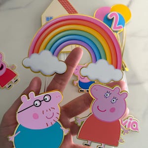 May include: A collection of Peppa Pig-themed birthday cake toppers. The toppers include a rainbow, clouds, balloons, the number 2, and Peppa Pig characters. The characters are outlined in gold and have a cartoon style.