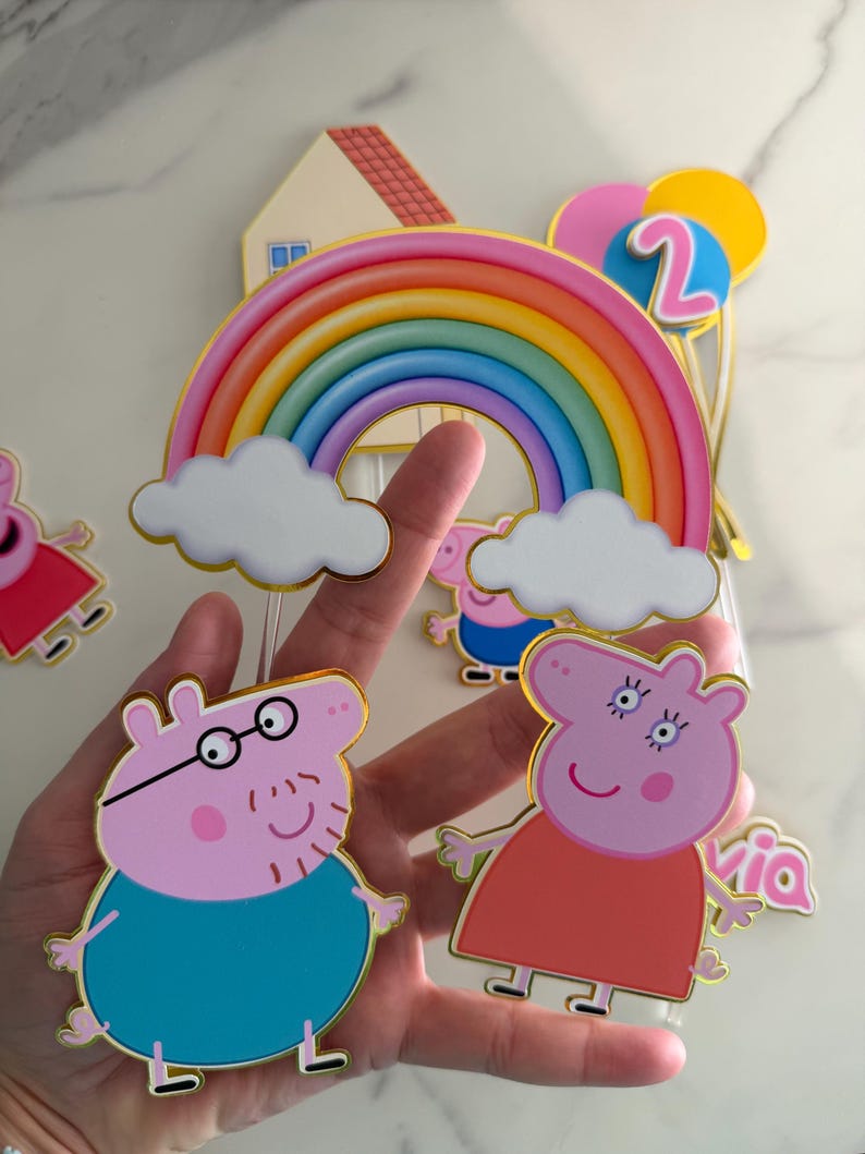 May include: A collection of Peppa Pig-themed birthday cake toppers. The toppers include a rainbow, clouds, balloons, the number 2, and Peppa Pig characters. The characters are outlined in gold and have a cartoon style.