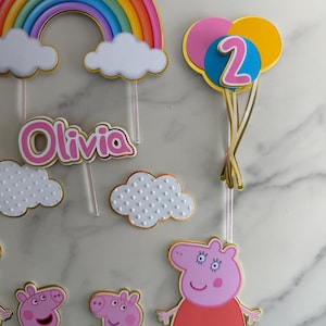 May include: Cake toppers featuring a rainbow, clouds, the name "Olivia", and Peppa Pig characters. A balloon cluster with the number "2" is also included. The toppers are in various colors with gold accents, perfect for a birthday celebration.