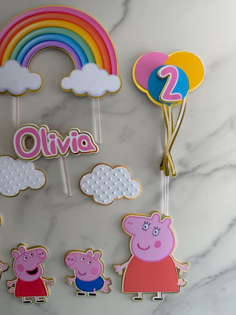 May include: Cake toppers featuring a rainbow, clouds, the name "Olivia", and Peppa Pig characters. A balloon cluster with the number "2" is also included. The toppers are in various colors with gold accents, perfect for a birthday celebration.