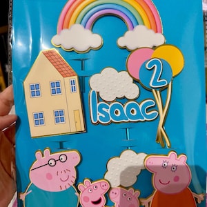 May include: Cake toppers featuring a rainbow, clouds, a house, balloons with the number 2, and the name "Isaac". The toppers also include cartoon pig characters. The background is a blue plastic sheet.
