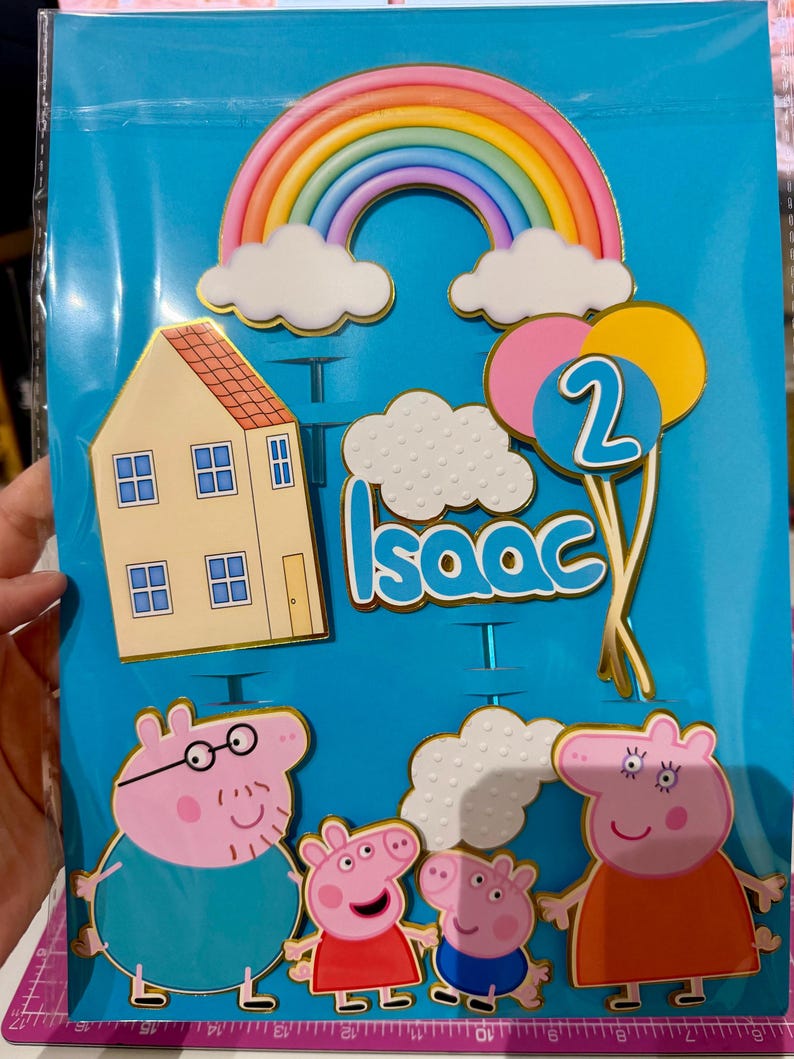 May include: Cake toppers featuring a rainbow, clouds, a house, balloons with the number 2, and the name "Isaac". The toppers also include cartoon pig characters. The background is a blue plastic sheet.