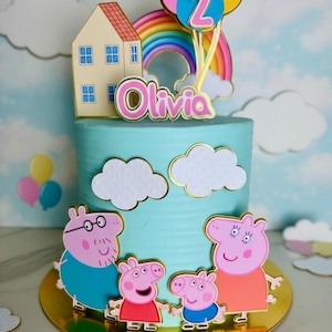May include: A birthday cake with blue frosting, decorated with cartoon characters, a rainbow, and balloons. The cake has a gold base and is topped with a house and the name "Olivia". The number "2" is on the balloons.
