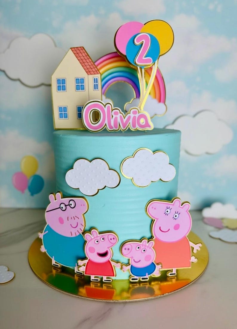 May include: A birthday cake with blue frosting, decorated with cartoon characters, a rainbow, and balloons. The cake has a gold base and is topped with a house and the name "Olivia". The number "2" is on the balloons.
