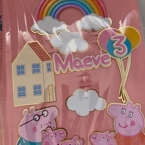 May include: A set of birthday cake toppers featuring a rainbow, clouds, a house, balloons, and Peppa Pig characters. The name "Maeve" and the number "3" are also included. The toppers are in a pink package.