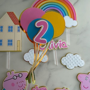 May include: A colorful birthday cake topper set featuring a rainbow, clouds, and balloons with the number 2. Includes Peppa Pig characters and the name "Olivia". Gold trim accents the decorations.