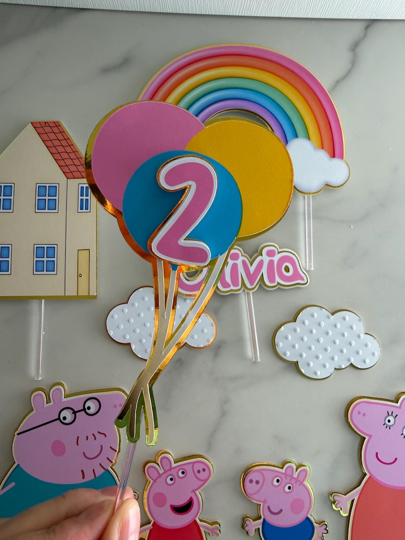 May include: A colorful birthday cake topper set featuring a rainbow, clouds, and balloons with the number 2. Includes Peppa Pig characters and the name "Olivia". Gold trim accents the decorations.