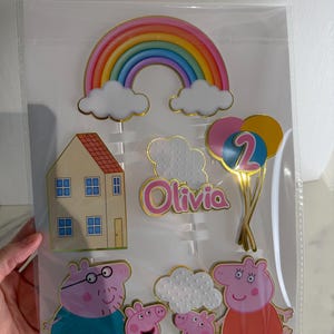 May include: Cake toppers featuring a rainbow, clouds, a house, balloons with the number 2, and a family of cartoon pigs. The name "Olivia" is also included. The toppers are in a clear plastic package.