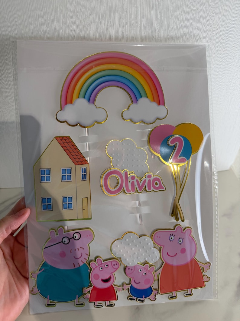 May include: Cake toppers featuring a rainbow, clouds, a house, balloons with the number 2, and a family of cartoon pigs. The name "Olivia" is also included. The toppers are in a clear plastic package.