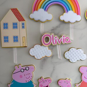 May include: Cake toppers featuring a rainbow, clouds, a house, and cartoon pig characters. The name "Olivia" is displayed in pink lettering. The toppers have gold trim and are designed for a birthday cake.