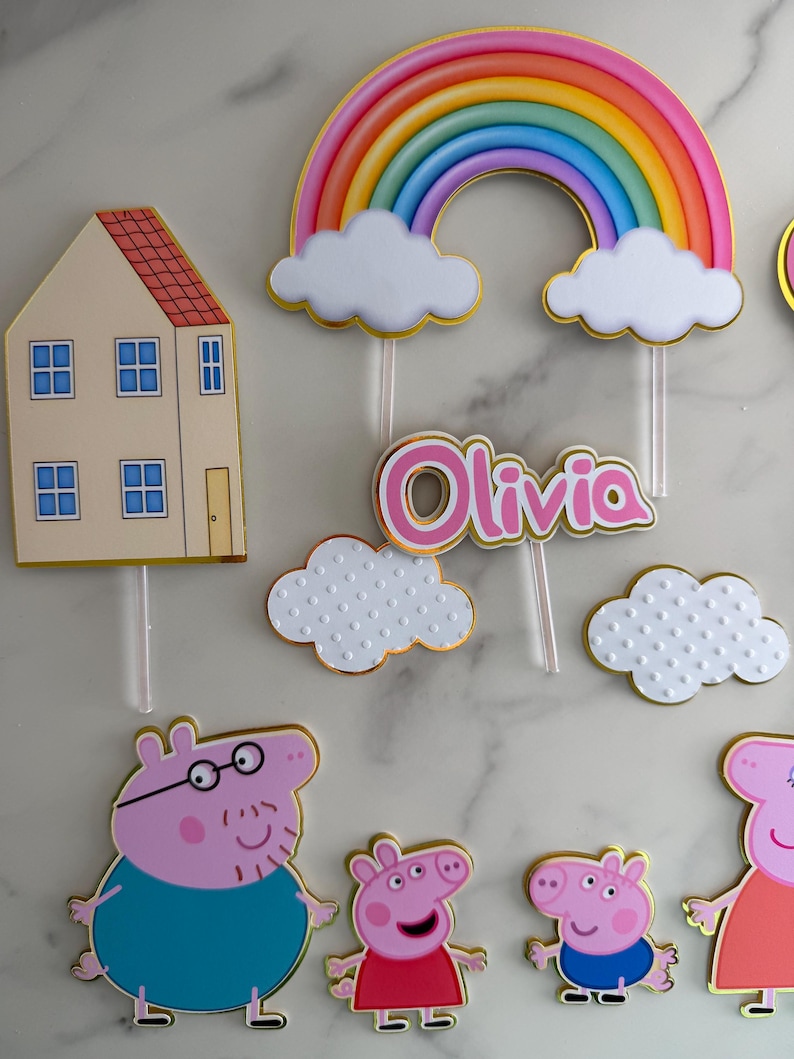May include: Cake toppers featuring a rainbow, clouds, a house, and cartoon pig characters. The name "Olivia" is displayed in pink lettering. The toppers have gold trim and are designed for a birthday cake.