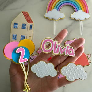 May include: A collection of colorful birthday cake toppers. Includes a rainbow, clouds, a house, balloons with the number 2, and a name topper that says "Olivia". The toppers have gold accents and are made of cardstock.