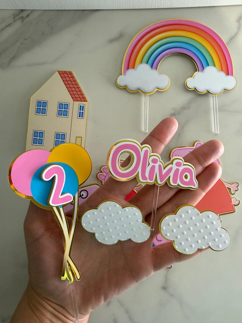 May include: A collection of colorful birthday cake toppers. Includes a rainbow, clouds, a house, balloons with the number 2, and a name topper that says "Olivia". The toppers have gold accents and are made of cardstock.