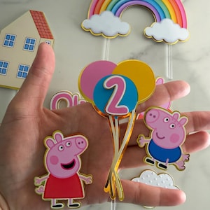 May include: A collection of colorful birthday decorations featuring a rainbow, clouds, and cartoon pig characters. Includes a house, balloons with the number 2, and pig figures in red and blue outfits. Perfect for a child's birthday party.