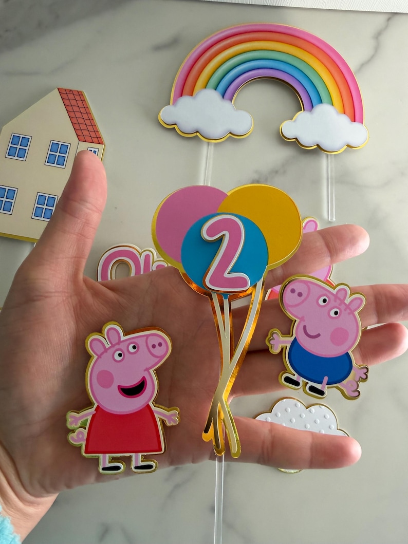 May include: A collection of colorful birthday decorations featuring a rainbow, clouds, and cartoon pig characters. Includes a house, balloons with the number 2, and pig figures in red and blue outfits. Perfect for a child's birthday party.