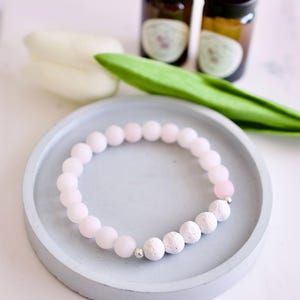 May include: A pink and white beaded bracelet with lava stone beads. The bracelet is on a grey concrete dish.
