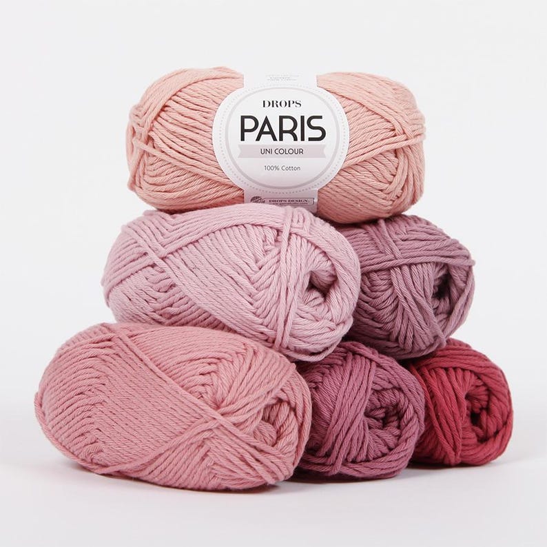 May include: A stack of Drops Paris Uni Colour cotton yarn balls in various shades of pink. The top ball has a label that reads "Drops Paris Uni Colour 100% Cotton."