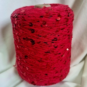 May include: A bright red yarn spool with embedded sequins. The yarn is wound around a cardboard core, forming a cylindrical shape. The sequins are scattered across the yarn's surface, adding a decorative element. Ideal for knitting and other craft projects.