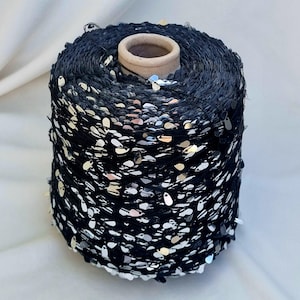 Könnte beinhalten: Eine Spule schwarzes Garn, dicht mit silbernen Pailletten bedeckt. Das Garn ist um einen Pappkern gewickelt. Die Pailletten sind unregelmäßig verteilt und erzeugen einen funkelnden Effekt. Ideal für Bastelarbeiten und Verzierungen.