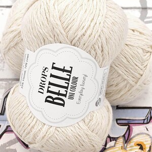 以下が含まれることがあります： 「DROPS BELLE UNI COLOUR Everyday luxury!」と書かれたラベルが付いた、クリーム色の毛糸玉のクローズアップ。 この糸はビスコースとリネンの混合で、編み物や編み物プロジェクトに最適です。