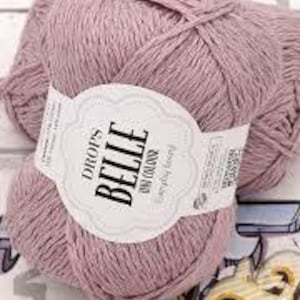 以下が含まれることがあります： ダスティローズ色のDrops Belleの糸玉2つのクローズアップ。糸は、装飾的なフォントで「DROPS BELLE」と「uni color」と書かれた白いラベルで包まれています。糸は白とグレーの模様の表面にあります。
