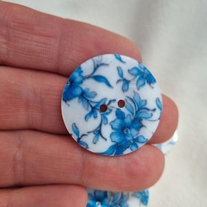 Può includere: Un bottone rotondo bianco con un motivo floreale blu. Il bottone ha due fori e una finitura lucida. Il motivo floreale blu presenta fiori e foglie dettagliate, ideale per cucito e artigianato.