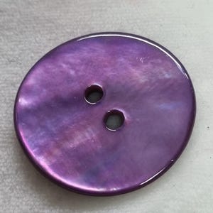 Op de afbeelding: Een grote, ovale knoop met een glanzende, iriserende paarse afwerking. De knoop heeft twee kleine gaatjes om aan te naaien. Het oppervlak van de knoop reflecteert licht en toont een reeks paarse tinten.
