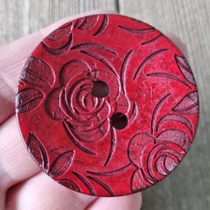 Könnte beinhalten: Ein großer, runder, roter Knopf mit floralem Design. Der Knopf zeigt geschnitzte Rosen und Blätter mit zwei Löchern zum Nähen. Der Knopf besteht aus einem festen Material und hat ein leichtes Used-Aussehen.