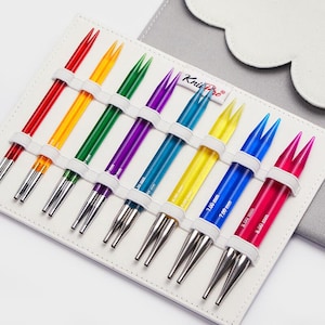 Peut inclure: Un ensemble d'aiguilles à tricoter colorées dans un étui blanc. Les aiguilles, aux couleurs rouge, jaune, vert, violet, bleu et rose, sont maintenues par des bandes élastiques blanches. Les pointes sont argentées.