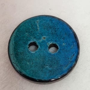 Op de afbeelding: Een ronde knoop met twee gaten en een kleurverloop van diep petrol naar helderblauw. Het oppervlak heeft een getextureerde, licht verweerde afwerking met zichtbare fijne barstjes en een donkere, gladde buitenrand rondom de cirkelvorm.