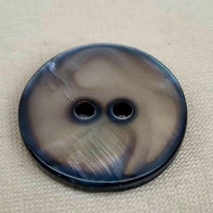 Puede incluir: Un botón grande y redondo con dos agujeros. El botón tiene un acabado nacarado e iridiscente con tonos grises, marrones y azules. El botón probablemente está hecho de concha o un material similar.