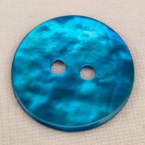 May include: A round, iridescent button with a teal and blue color scheme. The button has two holes and a glossy finish. The button is suitable for sewing projects and crafts.
