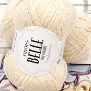 以下が含まれることがあります： 「DROPS BELLE UNI COLOURS Everyday Luxury」と書かれたラベルが付いた、クリーム色の毛糸玉。明るい木目調の背景に、模様のある表面に毛糸が展示されています。