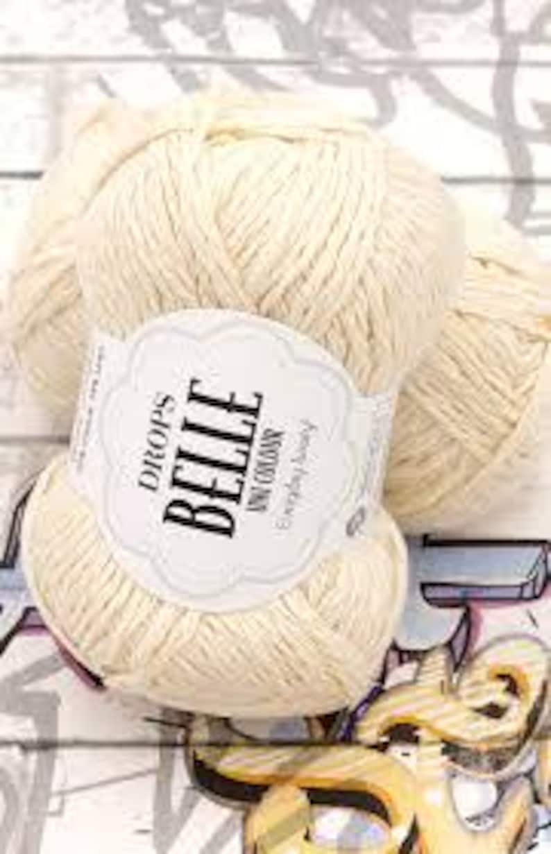 以下が含まれることがあります： 「DROPS BELLE UNI COLOURS Everyday Luxury」と書かれたラベルが付いた、クリーム色の毛糸玉。明るい木目調の背景に、模様のある表面に毛糸が展示されています。