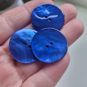 May include: Three round, blue buttons with a pearlescent sheen. Each button has two small holes for sewing. The buttons are a deep, rich blue color, with a swirling, iridescent effect.