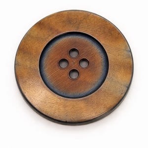 Könnte beinhalten: Ein großer, runder Knopf mit metallischem Finish. Er hat einen zentralen, vertieften Bereich mit vier Nählöchern. Der äußere Rand ist bronzefarben, während die Mitte einen dunkleren, kupferfarbenen Farbton aufweist. Durchmesser ca. 4 cm.