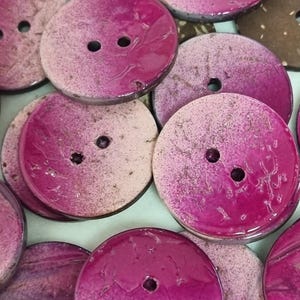 Könnte beinhalten: Eine Sammlung runder Knöpfe mit zwei Löchern und einem Farbverlauf, der von einem tiefen, leuchtenden Magenta zu einem hellen Rosa übergeht. Die Oberfläche jedes Knopfes zeigt eine natürliche, texturierte Maserung mit einer glatten, glänzenden Schicht.