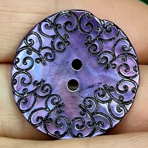 Può includere: Un bottone rotondo, viola iridescente con due fori. Il bottone presenta un design a filigrana nero e ornato con volute sui bordi. Il bottone è tenuto tra due dita.
