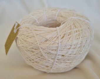 May include: A ball of off-white yarn with a small, gold-colored tag attached. The yarn is tightly wound, and white thread is wrapped around the ball. The yarn appears to be made of natural fibers, suitable for knitting or crocheting projects.