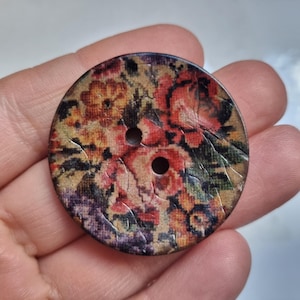 Op de afbeelding: Een grote, ronde knoop met een bloemenpatroon. De knoop heeft een patroon van rode, oranje en paarse bloemen op een beige achtergrond. Het heeft twee gaten om te naaien en een licht verweerde uitstraling.