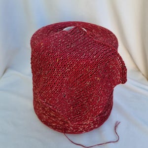 May include: A large spool of red yarn with gold metallic threads woven throughout. The yarn is partially unraveled, revealing the knitted texture. The yarn is suitable for knitting or crocheting projects.