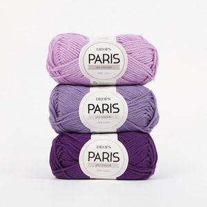 May include: Three balls of Drops Paris Uni Colour yarn in shades of purple, stacked on top of each other. Each ball has a white label with the brand name and product details. The yarn is 100% cotton.