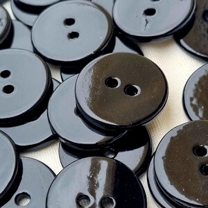 Puede incluir: Un conjunto de botones redondos negros brillantes, cada uno con dos agujeros. Los botones están dispersos, algunos se superponen, creando una superficie texturizada. Los botones tienen un brillo reflectante, lo que sugiere un acabado liso. Ideal para coser y manualidades.