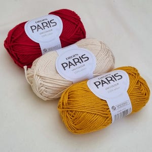 May include: Three skeins of Drops Paris Uni Colour 100% cotton yarn in red, cream, and mustard yellow. Each skein has a label with the brand name and fiber content. The yarn is suitable for various knitting and crochet projects.