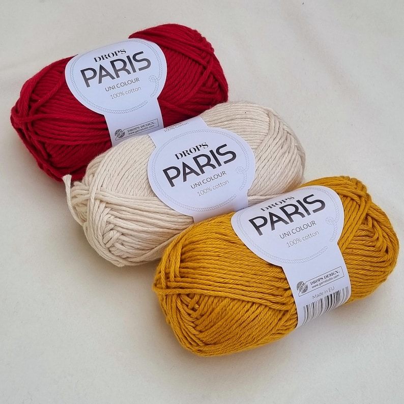 May include: Three skeins of Drops Paris Uni Colour 100% cotton yarn in red, cream, and mustard yellow. Each skein has a label with the brand name and fiber content. The yarn is suitable for various knitting and crochet projects.