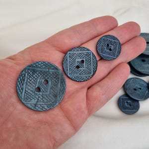 Può includere: Una collezione di bottoni blu-grigi di varie dimensioni. Ogni bottone presenta un design quadrato all'interno di una forma circolare, con un motivo a tratteggio incrociato e due fori per cucire. Alcuni bottoni sono in una mano.