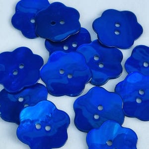Può includere: Una collezione di bottoni blu vibranti a forma di fiore. Ogni bottone ha due fori per cucire e una finitura lucida. I bottoni sono sparsi su una superficie bianca, creando una disposizione visivamente accattivante. Ideali per cucito e progetti artigianali.