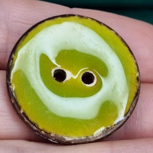 Peut inclure: Un bouton en céramique vert et jaune avec deux trous. Le bouton a un motif en spirale blanc.